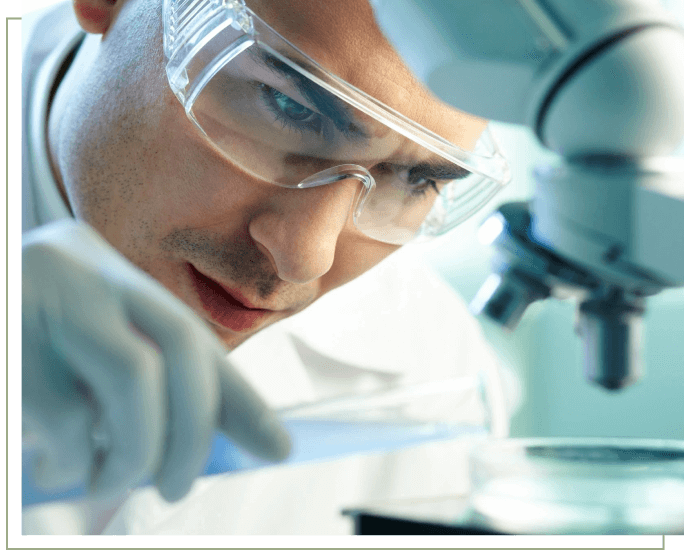 A man in lab coat and goggles looking at a microscope.